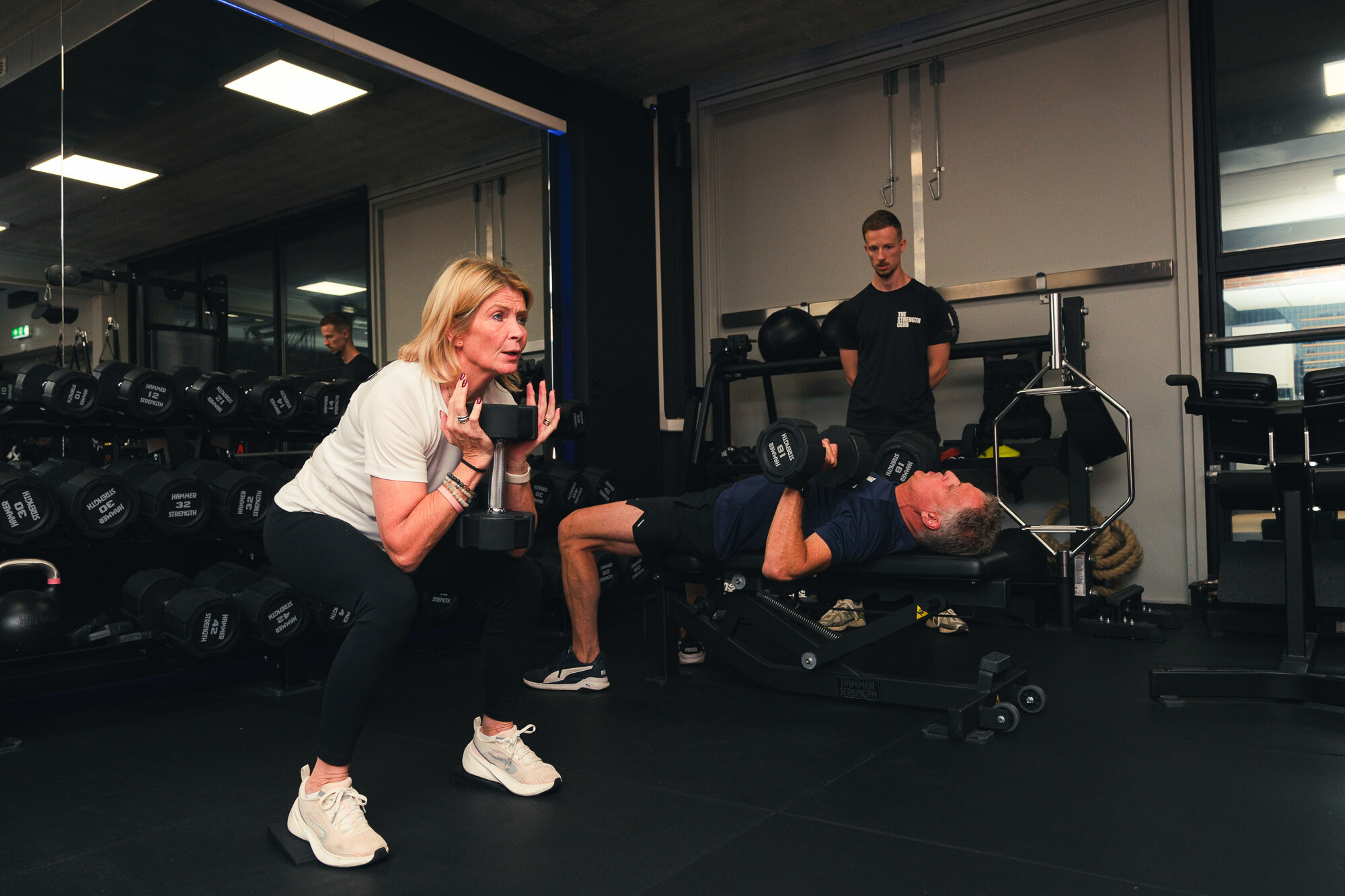 Twee sporters doen krachttraining met dumbbells onder begeleiding van een personal trainer