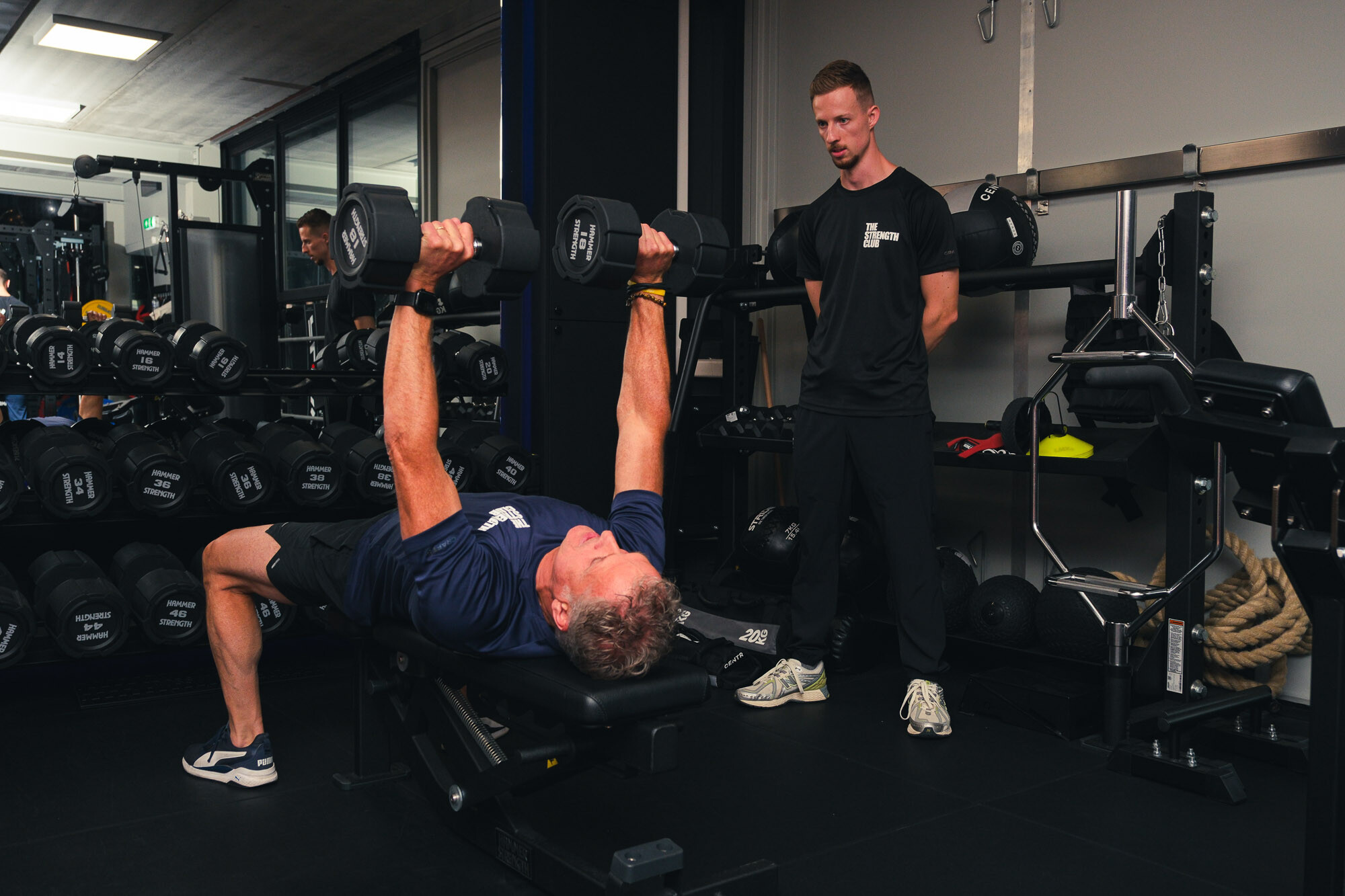 Sporter doet bankdrukken met dumbbells onder begeleiding