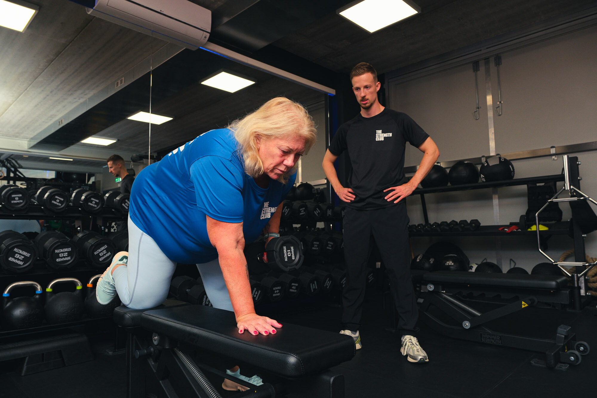 Sporter doet een dumbbell row op een trainingsbank onder begeleiding Sporter doet een dumbbell row op een trainingsbank onder begeleiding