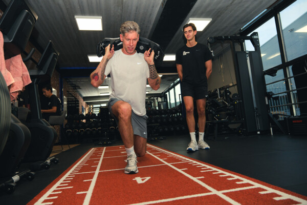 Sporter doet een lunge met gewichten op schouderhoogte onder begeleiding Sporter doet een lunge met gewichten op schouderhoogte onder begeleiding