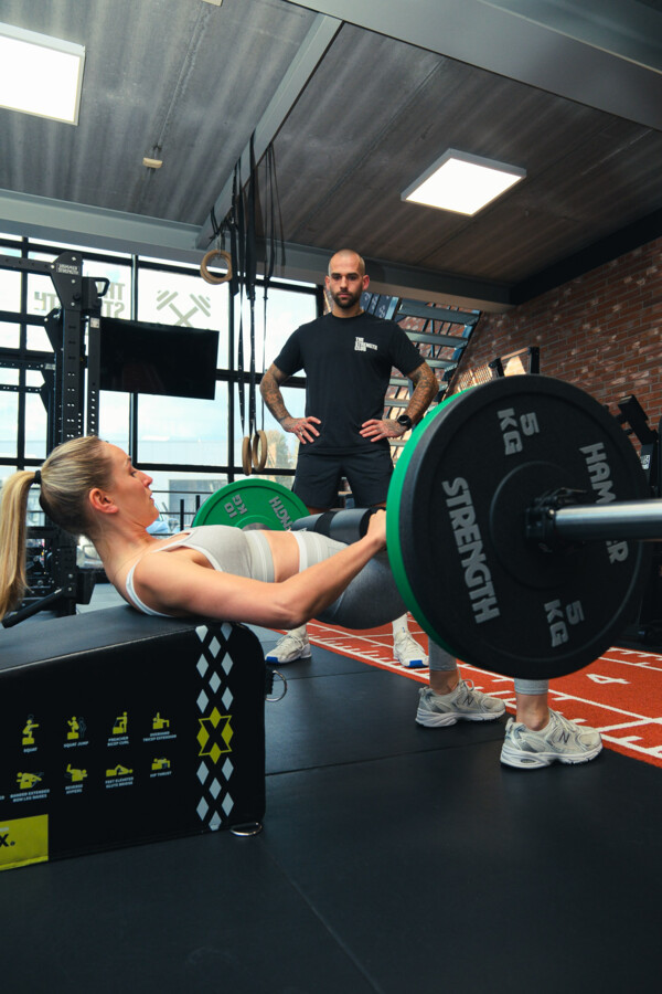 Personal trainer begeleidt een sporter bij een hip thrust met halterstang