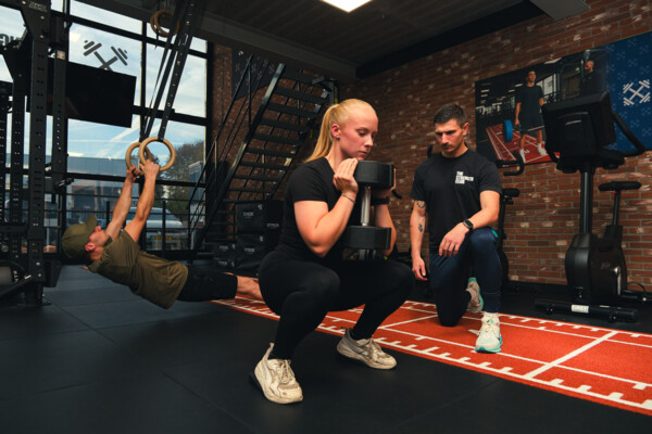 Sporter doet een goblet squat terwijl een andere sporter aan gymnastiekringen traint Sporter doet een goblet squat terwijl een andere sporter aan gymnastiekringen traint